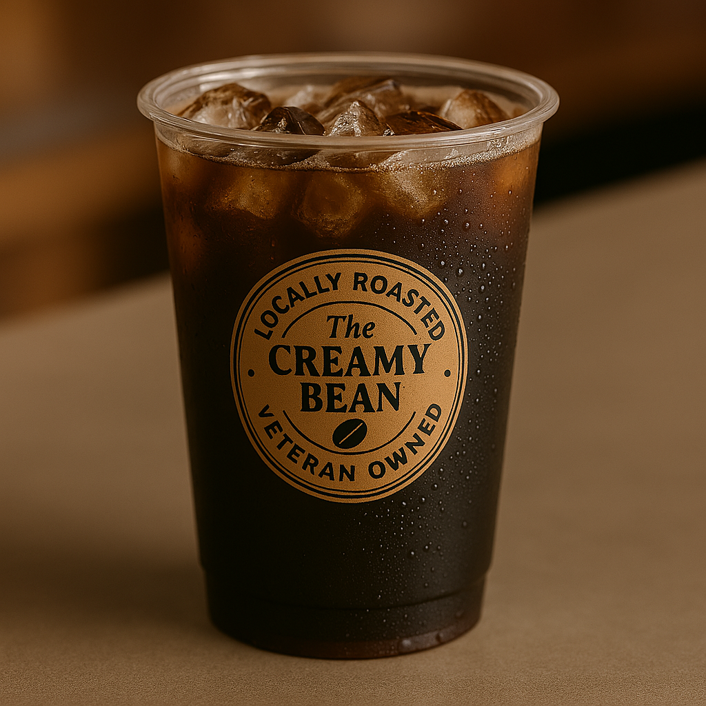 A clear plastic to-go cup without a lid, filled with a 12oz iced Americano — bold black espresso over ice, featuring The Creamy Bean logo, set against a tropical background with palm trees and a warm orange-to-teal gradient.