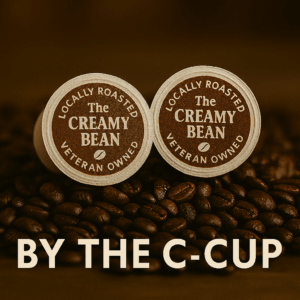 Creamy Bean C-Cup coffee pods sitting on roasted coffee beans with logo on top and "BY THE C-CUP" text along the bottom.