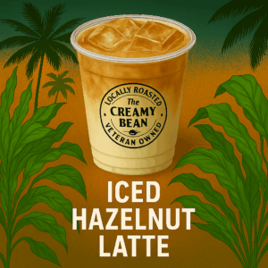 A clear plastic to-go cup without a lid, filled with a creamy iced hazelnut latte and visible ice cubes. The Creamy Bean logo is printed on the cup. The background features tropical palm trees and lush green leaves with a warm orange-to-teal gradient sunrise. “ICED HAZELNUT LATTE” is displayed in bold white capital letters along the bottom.